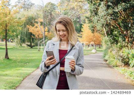 Smiling young woman using her phone while walking in autumn park for chatting, shopping, remote work, content creation for social media. Girl holding reusable cup with hot drink. and checking massages Smiling young woman using her phone while walking in autumn park for chatting, shopping, remote work, content creation for social media. Girl holding reusable cup with hot drink. and checking massages 142489469