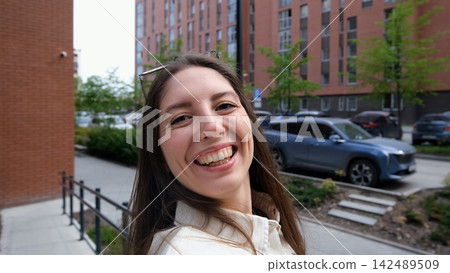 Smiling young woman taking selfie on city steps with modern apartment buildings behind her, casual white shirt, relaxed urban lifestyle, overcast daylight, close portrait view Smiling young woman taking selfie on city steps with modern apartment buildings behind her, casual white shirt, relaxed urban lifestyle, overcast daylight, close portrait view 142489509