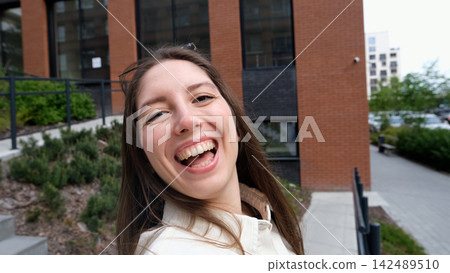 Smiling young woman taking selfie on city steps with modern apartment buildings behind her, casual white shirt, relaxed urban lifestyle, overcast daylight, close portrait view 142489510