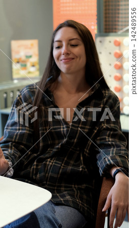 Smiling young woman in plaid shirt sitting at table in casual indoor setting, relaxed posture, soft natural light, long dark hair, comfortable modern workspace or cafe atmosphere Smiling young woman in plaid shirt sitting at table in casual indoor setting, relaxed posture, soft natural light, long dark hair, comfortable modern workspace or cafe atmosphere 142489556