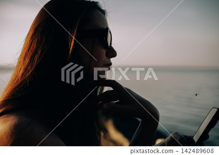 Woman in sunglasses sitting beside the sea at golden hour, looking at a smartphone. Warm backlight and calm water create a moody coastal summer lifestyle scene about travel, solitude and digital life. Woman in sunglasses sitting beside the sea at golden hour, looking at a smartphone. Warm backlight and calm water create a moody coastal summer lifestyle scene about travel, solitude and digital life. 142489608