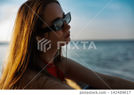 Woman wearing black sunglasses and red top by the sea at golden hour, with long hair illuminated by sunlight. Calm ocean and open sky create a stylish summer travel and lifestyle scene for modern ads. Woman wearing black sunglasses and red top by the sea at golden hour, with long hair illuminated by sunlight. Calm ocean and open sky create a stylish summer travel and lifestyle scene for modern ads. 142489617
