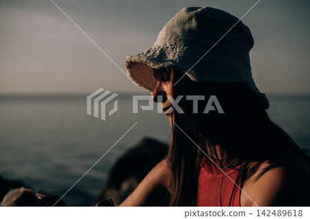 Woman in a white denim bucket hat and red sleeveless top sitting beside the sea at golden hour. Warm sunlight highlights her profile while the calm water and muted tones create a moody summer mood. 142489618