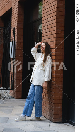 Smiling young woman leaning on brick pillar outdoors in casual white shirt, wide blue jeans, and sneakers, standing beside modern building entrance with relaxed confident pose 142489679