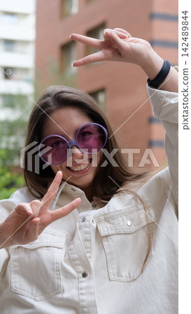 Cute cheerful charming girl poses for camera in summer on city street, hands and fingers showing V sign Cute cheerful charming girl poses for camera in summer on city street, hands and fingers showing V sign 142489844