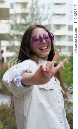 Cute cheerful charming girl poses for camera in summer on city street, hands and fingers showing V sign 142489845