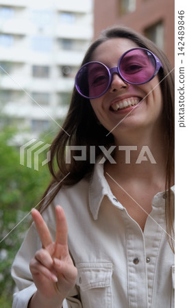 Cute cheerful charming girl poses for camera in summer on city street, hands and fingers showing V sign Cute cheerful charming girl poses for camera in summer on city street, hands and fingers showing V sign 142489846