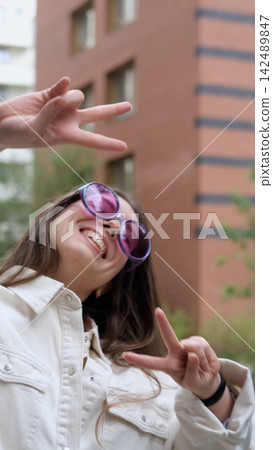 Cute cheerful charming girl poses for camera in summer on city street, hands and fingers showing V sign Cute cheerful charming girl poses for camera in summer on city street, hands and fingers showing V sign 142489847