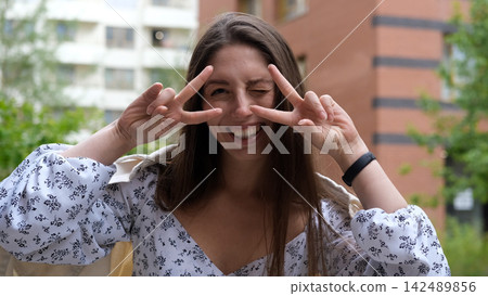 Cute cheerful charming girl poses for camera in summer on city street, hands and fingers showing V sign Cute cheerful charming girl poses for camera in summer on city street, hands and fingers showing V sign 142489856