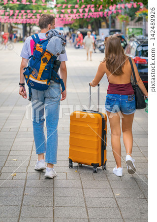 Back view of couple of travelers with suitcase walking through central Burgas during summer. Man carrying backpack and woman pulling orange suitcase walk along pedestrian street Back view of couple of travelers with suitcase walking through central Burgas during summer. Man carrying backpack and woman pulling orange suitcase walk along pedestrian street 142489926