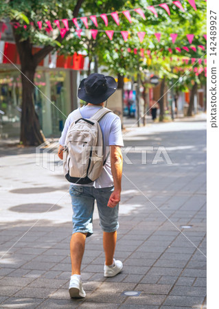 Man with Backpack Walking Along Pedestrian City Street. Rear view of casually dressed man with backpack walking along sunny pedestrian street. Urban solo travel, sightseeing, vacation, city lifestyle Man with Backpack Walking Along Pedestrian City Street. Rear view of casually dressed man with backpack walking along sunny pedestrian street. Urban solo travel, sightseeing, vacation, city lifestyle 142489927