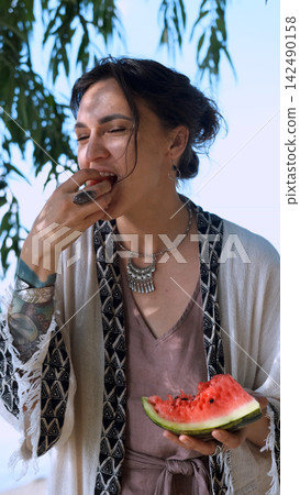 A beautiful brunette in a bohemian style relaxes outdoors in the summer, holding a large slice of watermelon against a green forest backdrop. A beautiful brunette in a bohemian style relaxes outdoors in the summer, holding a large slice of watermelon against a green forest backdrop. 142490158