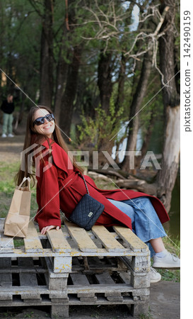 Portrait photo of beautiful young smiling woman in burgundy coat sitting on bench outdoors in park. Portrait photo of beautiful young smiling woman in burgundy coat sitting on bench outdoors in park. 142490159