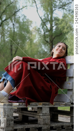 Portrait photo of beautiful young smiling woman in burgundy coat sitting on bench outdoors in park. Portrait photo of beautiful young smiling woman in burgundy coat sitting on bench outdoors in park. 142490163