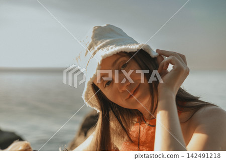Young woman in a white bucket hat and red top looking at the camera beside the sea. Warm sunlight and soft ocean background create a natural summer portrait with relaxed vacation mood and beauty chic. Young woman in a white bucket hat and red top looking at the camera beside the sea. Warm sunlight and soft ocean background create a natural summer portrait with relaxed vacation mood and beauty chic. 142491218