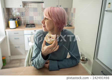 Smiling mature woman enjoys bitten donut after returning to sweets during PMS sugar craving. Smiling mature woman enjoys bitten donut after returning to sweets during PMS sugar craving. 142491498