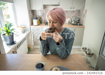 Mature woman bites chocolate donut during PMS craving and relapse into sugar addiction at home. Mature woman bites chocolate donut during PMS craving and relapse into sugar addiction at home. 142491518