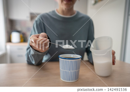 Woman adds white sugar to coffee with spoon, illustrating daily sugar dependence at home. Woman adds white sugar to coffee with spoon, illustrating daily sugar dependence at home. 142491536