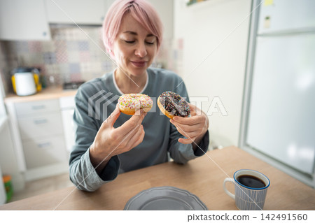 Smiling mature woman holds two donuts after giving in to sugar cravings during PMS at home. Smiling mature woman holds two donuts after giving in to sugar cravings during PMS at home. 142491560