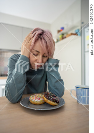 Mature woman struggles with PMS sugar cravings while resisting colorful donuts in home kitchen. Mature woman struggles with PMS sugar cravings while resisting colorful donuts in home kitchen. 142491570