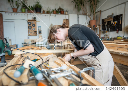 Concentrated woman restorer remodeling and remaking old wooden details for window frame in workshop Concentrated woman restorer remodeling and remaking old wooden details for window frame in workshop 142491603