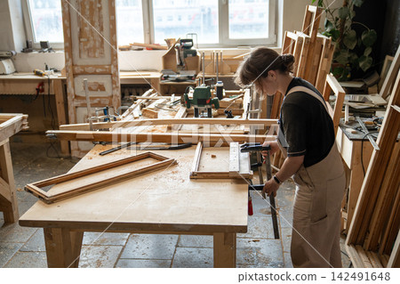Woman crafter in restoration workshop fixating parts of vintage wooden window frame using clamps. Woman crafter in restoration workshop fixating parts of vintage wooden window frame using clamps. 142491648