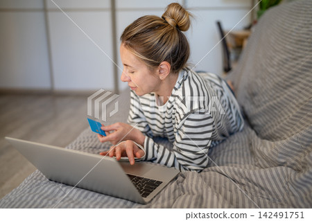 Carefree relaxed woman lying on sofa at home shopping online on laptop using bank credit card. Carefree relaxed woman lying on sofa at home shopping online on laptop using bank credit card. 142491751