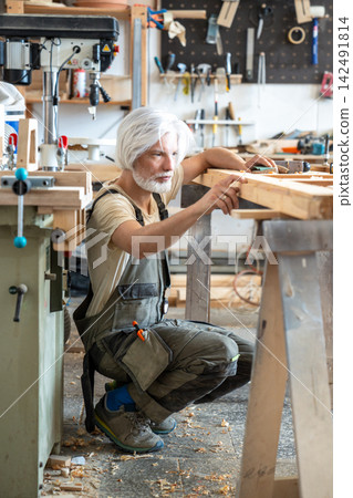 Passionate craftsman attentively checking surface of antique wooden door at worktable in workshop Passionate craftsman attentively checking surface of antique wooden door at worktable in workshop 142491814