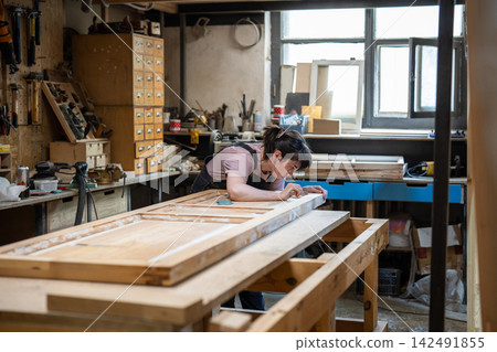 Passionate woman restorer carefully works on renewing old wooden door in restoration workshop. Passionate woman restorer carefully works on renewing old wooden door in restoration workshop. 142491855