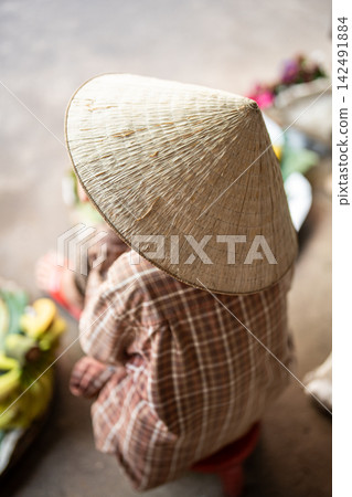 Unrecognizable street food market seller in traditional conical hat sitting waiting for customers. 142491884
