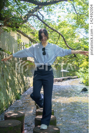 A Japanese woman in her 30s balances as she crosses a shallow, clear stream on stepping stones. 142492004