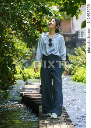 一位三十多歲的日本女子麵帶微笑地走在陽光斑駁的清澈溪流上的木棧道上。 142492006