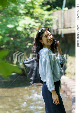 A woman in her 30s, carrying a shoulder bag, turns around and smiles on a riverside path. 142492017
