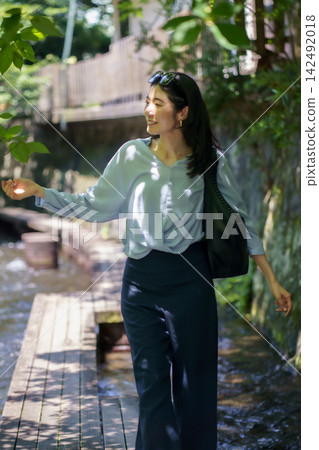 A Japanese woman in her 30s walks along a wooden boardwalk above a clear stream, offering her hand. A Japanese woman in her 30s walks along a wooden boardwalk above a clear stream, offering her hand. 142492018