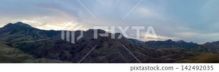 panoramic view of dark mountain ranges and undulating valleys beneath cloudy sunset sky panoramic view of dark mountain ranges and undulating valleys beneath cloudy sunset sky 142492035
