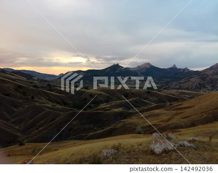 hilly mountain landscape with golden grass and rocky peaks in distance beneath evening clouds hilly mountain landscape with golden grass and rocky peaks in distance beneath evening clouds 142492036