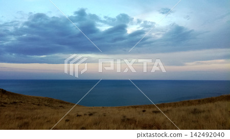 minimalist view of blue sea and dry grass hill beneath blue evening clouds minimalist view of blue sea and dry grass hill beneath blue evening clouds 142492040