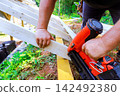 Builder works on construction project using pneumatic nail gun to secure wooden beams 142492380