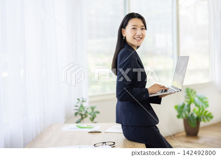 A young Japanese businesswoman smiles at the camera, holding a laptop, at a desk by a bright window. A young Japanese businesswoman smiles at the camera, holding a laptop, at a desk by a bright window. 142492800