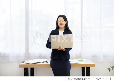 一位年輕的日本女商人面帶微笑地對著鏡頭,手裡拿著筆記型電腦,坐在明亮的窗邊的桌子旁。 一位年輕的日本女商人面帶微笑地對著鏡頭,手裡拿著筆記型電腦,坐在明亮的窗邊的桌子旁。 142492801