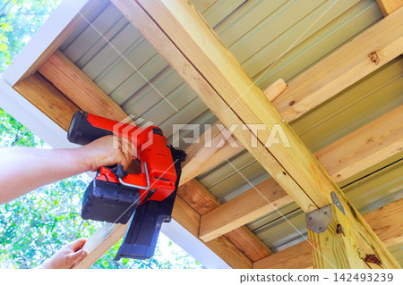 Contractor works on building project by using nail gun on wooden beams in yard 142493239