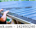 Builder works on installing metal roofing panels on structure while holding them in place. 142493240