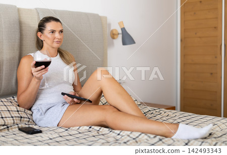 Gorgeous woman with glass of red wine relaxes lying on a bed in bedroom Gorgeous woman with glass of red wine relaxes lying on a bed in bedroom 142493343