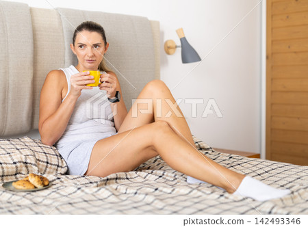 Adult woman drinking coffee sitting on bed in bedroom Adult woman drinking coffee sitting on bed in bedroom 142493346
