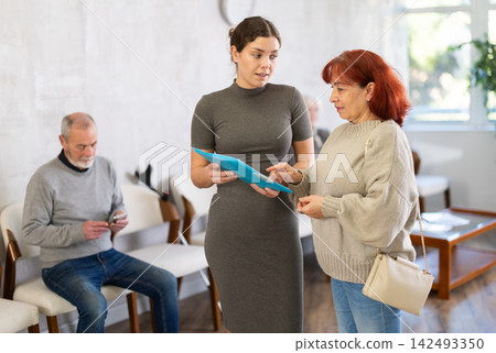 Positive polite nurse or doctor talking to aged man patient in reception area in background queue of people Positive polite nurse or doctor talking to aged man patient in reception area in background queue of people 142493350