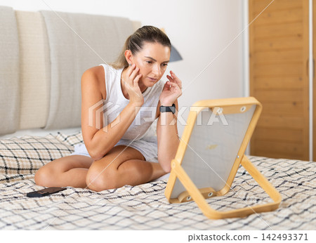 Woman in nightgown looks in the mirror and preens herself in interior of on bed in bedroom Woman in nightgown looks in the mirror and preens herself in interior of on bed in bedroom 142493371
