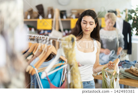 Young woman customer elect something summer clothes in store. Female client choosing something in background Young woman customer elect something summer clothes in store. Female client choosing something in background 142493381