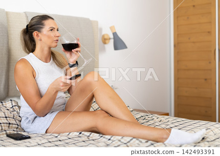 Woman relaxing at home in bedroom with glass of wine Woman relaxing at home in bedroom with glass of wine 142493391
