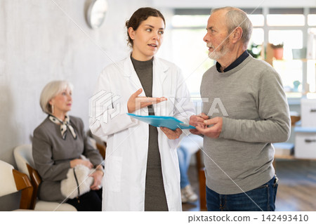 Polite young nurse or doctor talking to mature man patient standing in waiting room of medical clinic, with her back to visitors sitting in chairs Polite young nurse or doctor talking to mature man patient standing in waiting room of medical clinic, with her back to visitors sitting in chairs 142493410