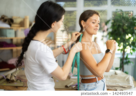 Female fashion designer taking body measurements of woman in tailoring studio Female fashion designer taking body measurements of woman in tailoring studio 142493547